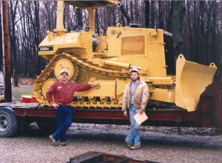 A new Cat D5H Dozer sitting on a trailer. This dozer was the first new piece of equipment purchased by Bob Simonson, pictured on the left next to the dozer operator, Jeff Moody.